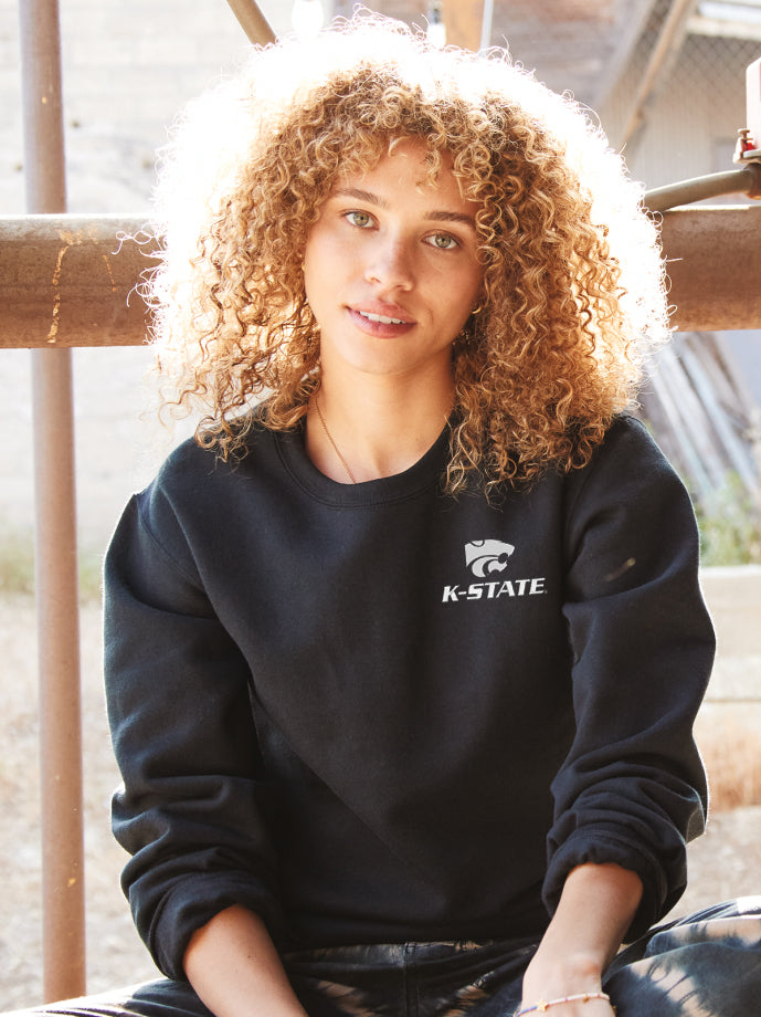 Female model, lifestyle shot, wearing black crewneck with K-state power cat logo on left chest.